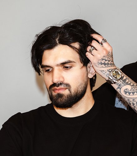 Man with tattoos and a beard touching his hair against a plain background